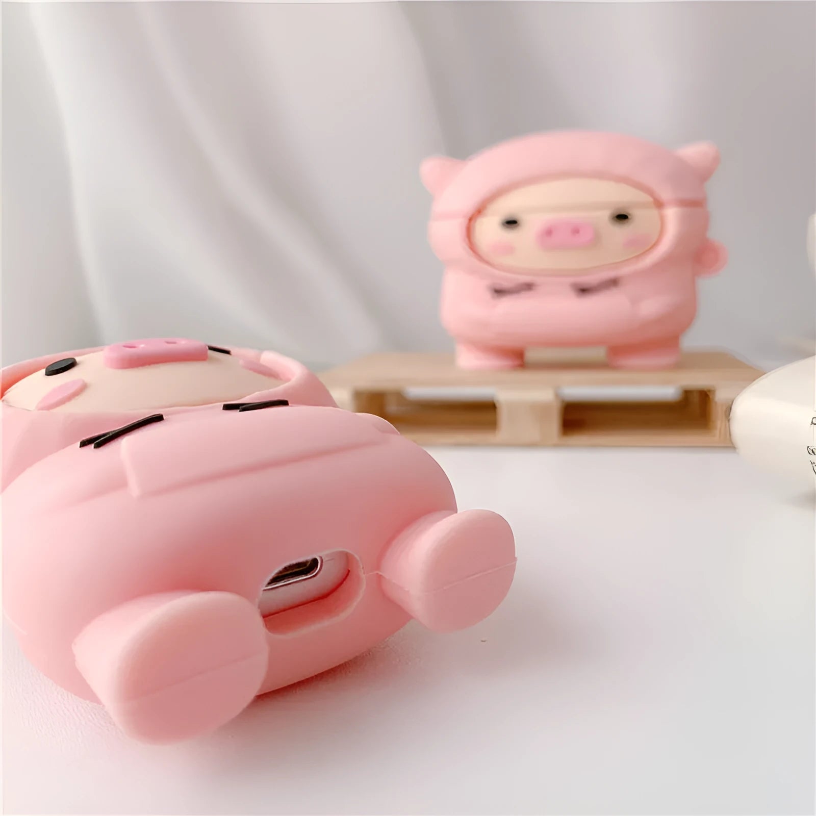Pink silicone AirPods case with cartoon pig in sweater design, shown with flip lid open and a matching metal hook.