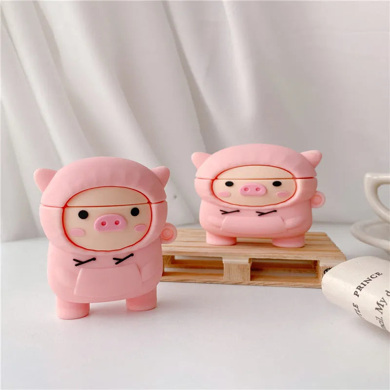 Pink silicone AirPods case with cartoon pig in sweater design, shown with flip lid open and a matching metal hook.