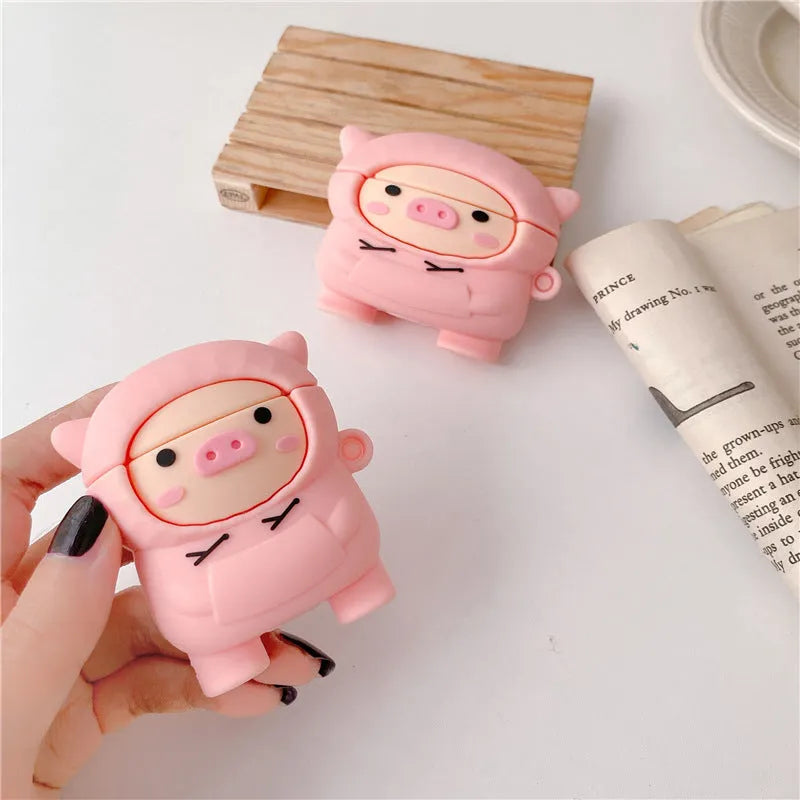 Pink silicone AirPods case with cartoon pig in sweater design, shown with flip lid open and a matching metal hook.