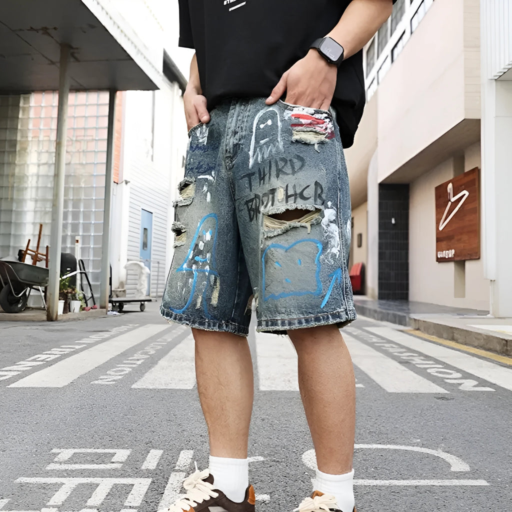 Blue men's denim jeans shorts with graffiti print and ripped details, showing straight leg and zipper fly.