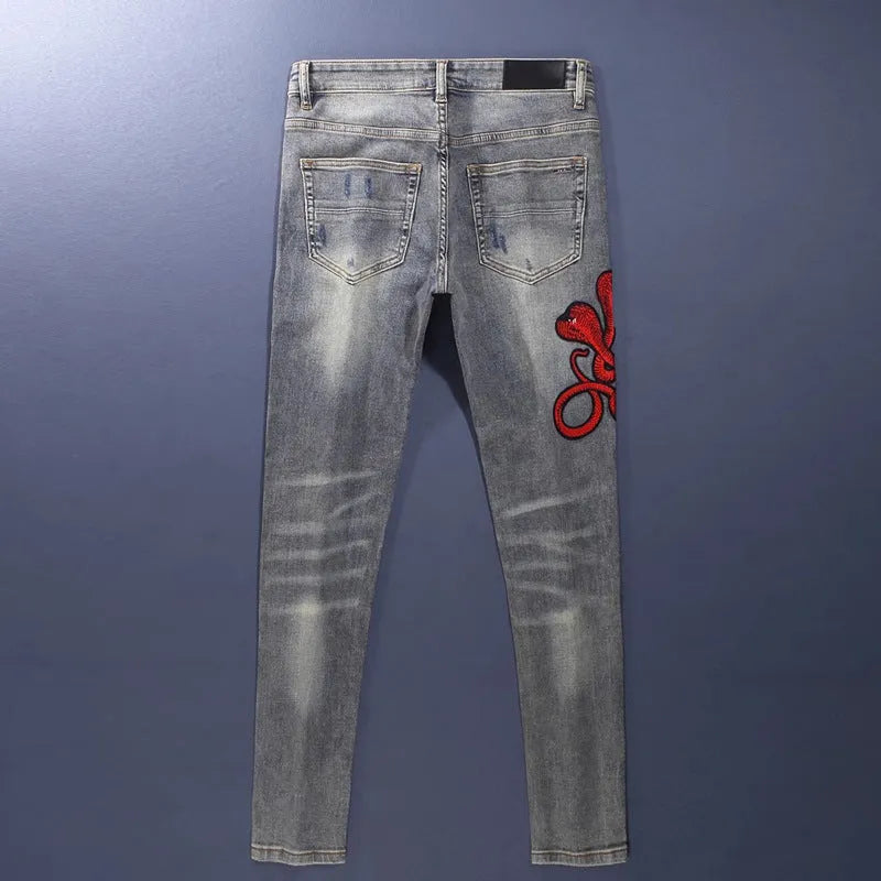 Man wearing slim fit distressed blue jeans with hole patches and embroidered details on the legs.