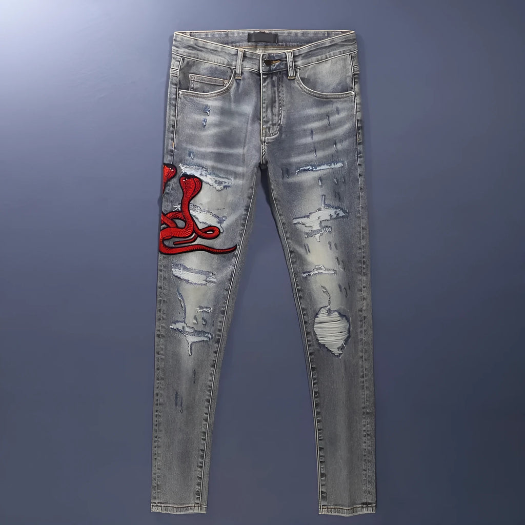 Man wearing slim fit distressed blue jeans with hole patches and embroidered details on the legs.